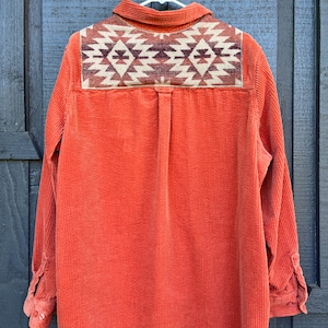 May include: An orange corduroy shirt with a geometric patterned yoke. The yoke features a design in shades of brown, cream, and burgundy. The shirt has long sleeves and buttoned cuffs, hanging on a white hanger.
