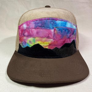 May include: A corduroy baseball cap with a brown brim and a colorful, patchwork design. The design features a sunset scene with a black mountain range at the bottom and a vibrant sky of pink, blue, and yellow hues.
