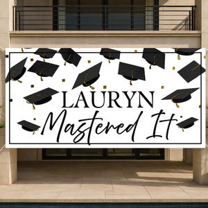 May include: A white banner with a black border, featuring the name "LAURYN" and the phrase "Mastered It" in a stylish script. Black graduation caps and gold squares are scattered across the banner, celebrating academic achievement.
