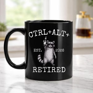 May include: Black ceramic mug with a raccoon illustration holding a bottle. The mug has the text "CTRL+ALT+ RETIRED EST. 2026" in white. The mug has a curved handle and is sitting on a white marble surface.