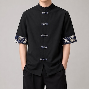 May include: Black short-sleeve shirt with a mandarin collar and decorative frog closures. The sleeves feature a navy blue and gold pattern with white crane illustrations. The shirt is made of a soft, comfortable material.