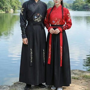 May include: Two traditional Chinese outfits. One is black with gold embroidery and a circular emblem with a bird. The other is red with black accents and gold embroidery. Both outfits have long skirts and are worn near a body of water.