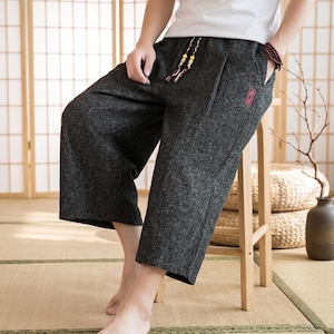 Puede incluir: Pantalones cortos de color gris carbón con cintura con cordón y bolsillos. Los pantalones tienen un aspecto texturizado y un pequeño diseño bordado en rojo cerca del bolsillo. El modelo lleva los pantalones sentado en un taburete de madera.