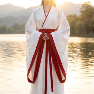 May include: A white traditional robe with long sleeves and a red sash. The robe has red trim on the sleeves and neckline. The garment is worn by a person standing in front of a lake and mountains.