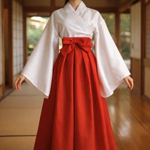Puede incluir: Un atuendo tradicional japonés hakama. El conjunto presenta una parte superior de kimono blanco con mangas anchas y un cierre envolvente. Un gran lazo rojo adorna la cintura, sobre una falda hakama roja plisada hasta el suelo. El atuendo está diseñado para ocasiones ceremoniales o formales.