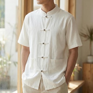 Puede incluir: Camisa blanca de manga corta con cuello mandarín y cuatro cierres de botones marrón oscuro. La camisa tiene dos bolsillos delanteros y está hecha de una tela ligera. El modelo lleva pantalones beige.