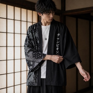 Puede incluir: Chaqueta negra estilo kimono con estampado de cachemira en el lado izquierdo y texto japonés en el lado derecho. La chaqueta se lleva sobre una camiseta blanca y pantalones negros. El texto japonés dice "Champion".