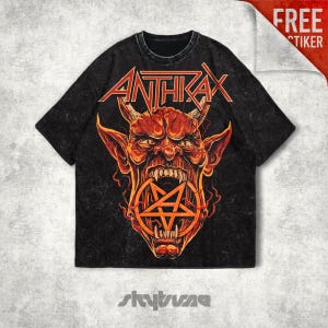 Puede incluir: Una camiseta negra con efecto lavado al ácido que presenta un gráfico de un rostro de demonio con cuernos en rojo y naranja con un símbolo de pentagrama en la boca. La palabra ANTHRAX está impresa en una fuente naranja sobre la ilustración.