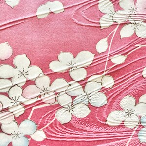 May include: A vibrant pink fabric featuring a repeating pattern of white cherry blossoms with delicate black stamen details. The background is textured with subtle, flowing horizontal lines that create a sense of movement across the surface.