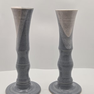 May include: Two ceramic candle holders. Each has a wide, circular base, a slender, textured stem, and a flared top. The colour scheme is a blend of grey and off-white, with a speckled appearance. The candle holders are displayed against a white background.