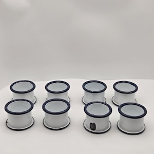 May include: A set of eight white enamel napkin rings, each with a dark blue trim. The napkin rings are cylindrical with a flat base and a dark blue rim. The set is displayed on a white surface.