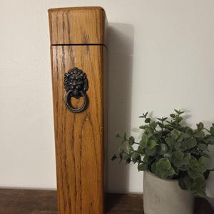 Peut inclure: Une boîte en bois rectangulaire et haute, ornée d'un heurtoir en forme de tête de lion. Le bois est brun clair avec des veines apparentes. La tête de lion est en métal foncé avec un anneau. Un petit pot de plante est à droite.