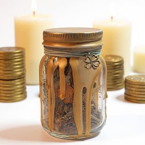 Puede incluir: Un tarro de cristal transparente lleno de hierbas y flores secas, coronado con una tapa dorada y un dije de tr&eacute;bol de cuatro hojas. Cera dorada gotea por el costado. El tarro est&aacute; rodeado de montones de monedas de oro y velas encendidas.