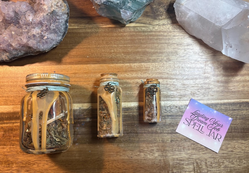 Puede incluir: Tres frascos de vidrio de diferentes tama&ntilde;os, llenos de hierbas secas y sellados con tapas decorativas. Los frascos est&aacute;n adornados con cera y cuerda. Tambi&eacute;n hay una peque&ntilde;a tarjeta con el texto "Landing Your Dream Job SPELL JAR".