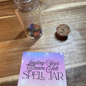 Puede incluir: Un tarro de cristal transparente lleno de piedras preciosas de colores, un tap&oacute;n de corcho y una tarjeta que dice "Landing Your Dream Job SPELL JAR" sobre una superficie de madera. El tarro es cil&iacute;ndrico.