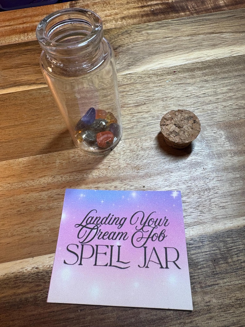 Puede incluir: Un tarro de cristal transparente lleno de piedras preciosas de colores, un tap&oacute;n de corcho y una tarjeta que dice "Landing Your Dream Job SPELL JAR" sobre una superficie de madera. El tarro es cil&iacute;ndrico.