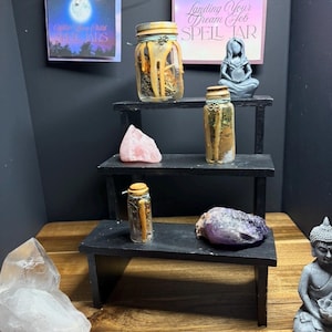 Puede incluir: Una exhibici&oacute;n de frascos de hechizos, cristales y una peque&ntilde;a estatua en un estante negro escalonado. Los frascos est&aacute;n llenos de hierbas y otros ingredientes. Dos letreros dicen "Landing Your Dream Job Spell Jar" y "Eight Moon Child Spell Jars".