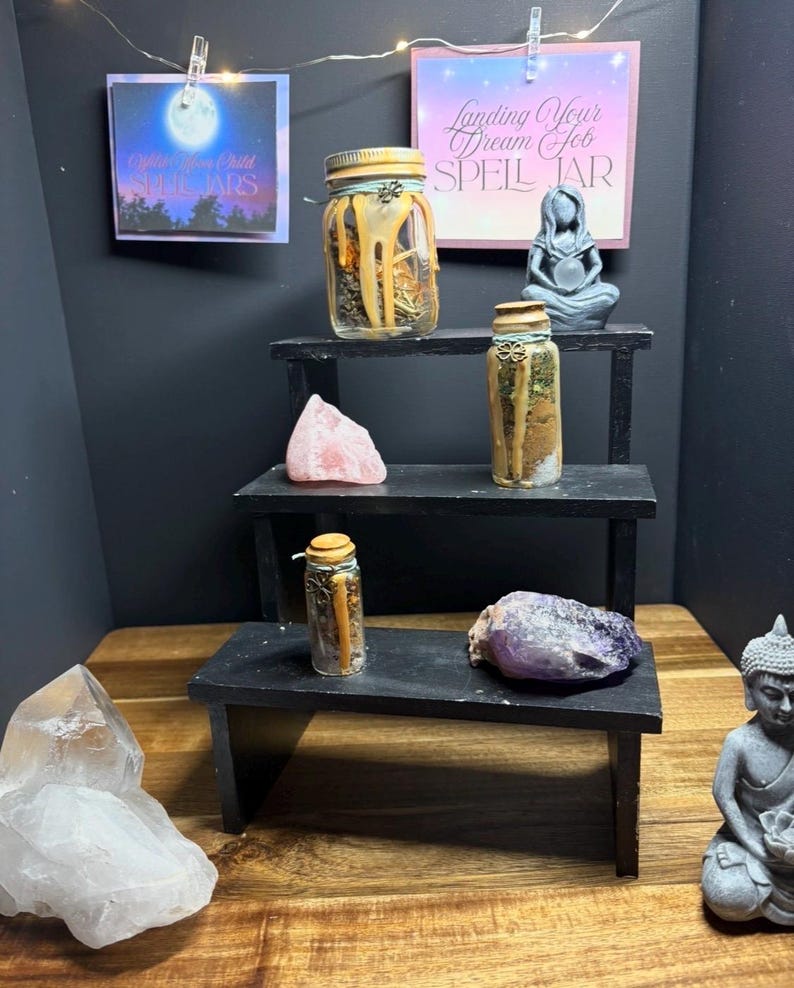 Puede incluir: Una exhibici&oacute;n de frascos de hechizos, cristales y una peque&ntilde;a estatua en un estante negro escalonado. Los frascos est&aacute;n llenos de hierbas y otros ingredientes. Dos letreros dicen "Landing Your Dream Job Spell Jar" y "Eight Moon Child Spell Jars".