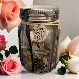 May include: A small glass jar with a silver lid and a decorative tag that reads "Never, never give up." The jar is filled with colorful dried herbs and crystals, and is decorated with black paint drips. The jar is tied with twine.