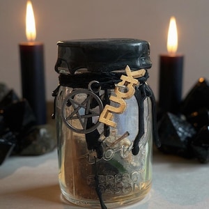 May include: A glass jar with a black lid and black wax drips, adorned with a pentagram charm and the word "FUCK" in gold letters. Two black candles and crystals flank the jar, with lit flames in the background.