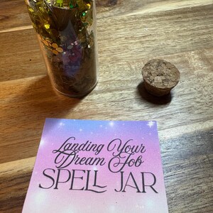 Puede incluir: Un tarro de cristal transparente lleno de purpurina dorada y verde y un tap&oacute;n de corcho. Una tarjeta con el texto "Landing Your Dream Job SPELL JAR" est&aacute; al lado del tarro. El fondo es una superficie de madera.