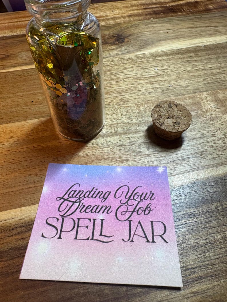 Puede incluir: Un tarro de cristal transparente lleno de purpurina dorada y verde y un tap&oacute;n de corcho. Una tarjeta con el texto "Landing Your Dream Job SPELL JAR" est&aacute; al lado del tarro. El fondo es una superficie de madera.