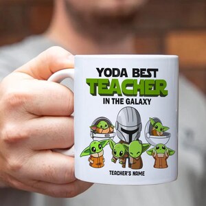 May include: White ceramic mug with the text "YODA BEST TEACHER IN THE GALAXY" in black and green. The mug features illustrations of Baby Yoda and a Mandalorian helmet. The bottom of the mug has space for a teacher's name.