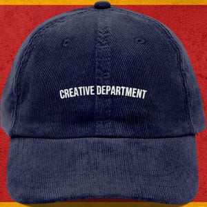 May include: A navy blue corduroy baseball cap with the words "CREATIVE DEPARTMENT" in white block letters across the front. The cap has a curved brim and a small button on top. The background is a red and yellow.