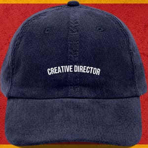 Może przedstawiać: Granatowa czapka bejsbolowa z sztruksu z białym napisem "CREATIVE DIRECTOR". Czapka ma zakrzywiony daszek i guzik na górze. Tło jest czerwone i żółte.