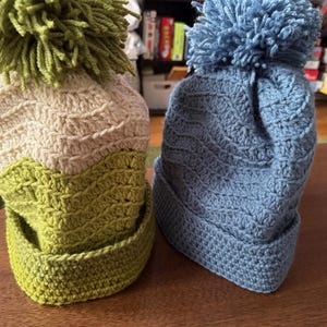 May include: Two handmade crochet beanies. One is green, cream, and light green with a green pom-pom. The other is blue with a blue pom-pom. Both beanies have a textured wave pattern and a folded brim.