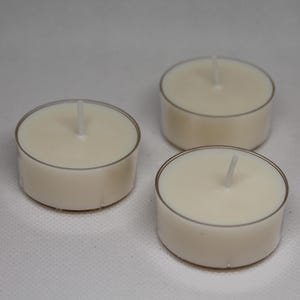 Puede incluir: Tres velas de té de color marfil con mechas blancas. Cada vela está colocada en una copa redonda de plástico transparente. Las velas están dispuestas sobre una superficie blanca.