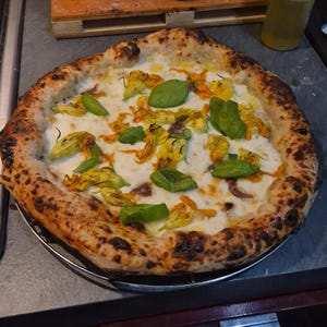 Può includere: Una pizza appena sfornata con una crosta dorata, condita con formaggio bianco, fiori di zucchina gialli e foglie di basilico verdi. La pizza è su una griglia di metallo, con vari barattoli e bottiglie sullo sfondo.
