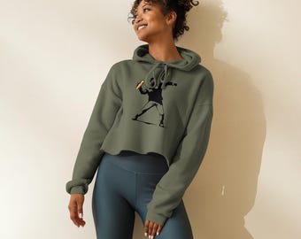 Op de afbeelding: Een olijfgroene cropped hoodie met een zwarte afbeelding van een persoon die een object gooit. De hoodie heeft een capuchon met trekkoord en lange mouwen. De persoon draagt ​​donkerblauwe fietsbroeken.