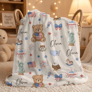 Puede incluir: Una manta blanca para bebé con ilustraciones patrióticas americanas, incluyendo un oso de peluche con un corazón de la bandera estadounidense, la Estatua de la Libertad y fuegos artificiales. El nombre "Olivia" está impreso.