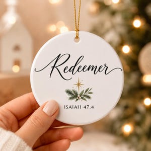 Peut inclure: Une décoration ronde en céramique blanche avec le mot Redeemer en écriture noire, une étoile dorée, des aiguilles de pin vertes et le texte Isaiah 47:4. L'ornement est suspendu par un fin cordon doré et possède un trou sur le dessus.