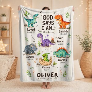 May include: A white blanket with a dinosaur theme. The blanket features colorful illustrations of various dinosaurs, each accompanied by a positive affirmation and a Bible verse. The name "OLIVER" is displayed at the bottom.