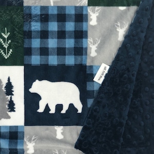 May include: A blue and gray patchwork baby blanket with a white bear, deer, and pine tree designs. The blanket has a soft, textured backing.