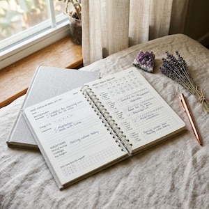 May include: A light grey journal, open to a page with a weekly mood tracker and daily check-in sections. A rose gold pen, lavender and a purple crystal are placed nearby. The journal is open to a page with handwritten notes and a spiral binding.