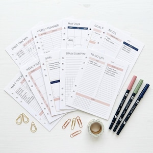 May include: A collection of planner inserts with headings like "Habit Tracker," "Weekly Planner," and "To-Do List." The inserts are white with pink and navy accents. Gold paper clips, washi tape, and three colored markers are also present.