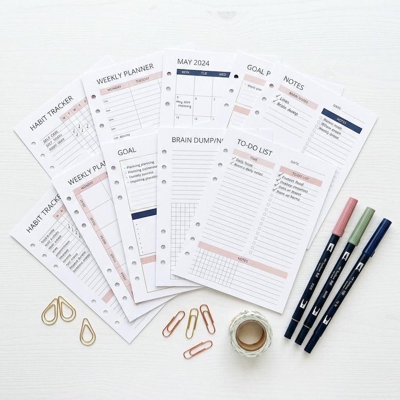 May include: A collection of planner inserts with headings like "Habit Tracker," "Weekly Planner," and "To-Do List." The inserts are white with pink and navy accents. Gold paper clips, washi tape, and three colored markers are also present.