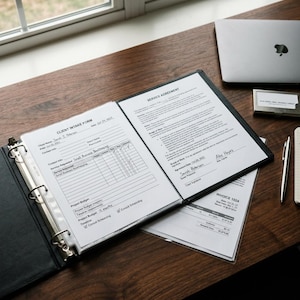 May include: An open black binder with documents, including a "Client Intake Form" and a "Service Agreement", lies on a wooden desk. A laptop, business card holder, and pen are also visible. The documents contain text and checkboxes.