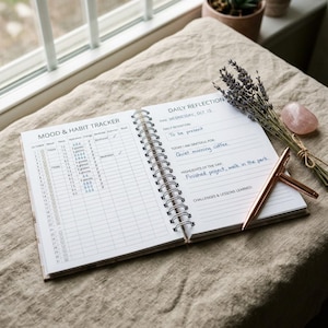 Puede incluir: Un diario abierto con espiral con el texto "MOOD & HABIT TRACKER" y "DAILY REFLECTION". Un bolígrafo de oro rosa descansa sobre la página, junto a un manojo de lavanda y un cristal rosa. El diario está sobre una superficie de lino beige.