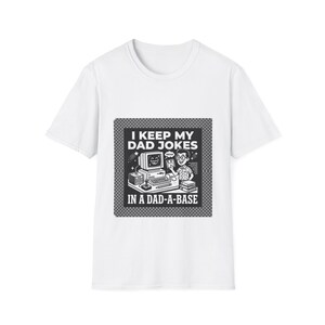 Può includere: T-shirt bianca con grafica in bianco e nero. La grafica presenta un computer retrò, una persona e il testo "I KEEP MY DAD JOKES IN A DAD-A-BASE."