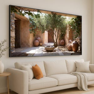 May include: A rectangular wall art print in a black frame depicting a sunlit Mediterranean courtyard. The scene features terracotta walls, a central olive tree, several large clay pots, a stone basin, and green vines hanging from an overhead structure.