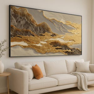 May include: A large framed artwork showing a stylized mountain landscape. The piece features textured layers in gold, grey, and beige, creating a three-dimensional effect. The artwork is displayed above a cream-coloured sofa.