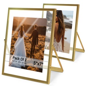 May include: Two gold-colored metal picture frames with clear glass, each holding a 5"x7" photograph. The frames have a simple, modern design with a stand on the back. One frame displays a wedding photo of a couple holding hands, and the other shows a woman on a beach.