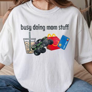 Puede incluir: Camiseta blanca con la frase "busy doing mom stuff" en letras negras. El diseño incluye una bebida, un monster truck, una caja roja de comida rápida y una tarjeta de crédito azul.