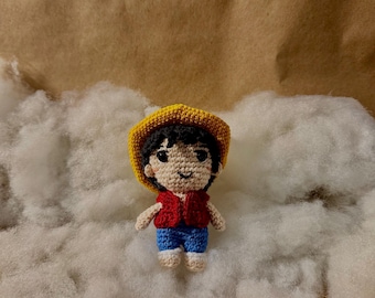May include: A crocheted doll of a cartoon character, wearing a yellow hat, red vest, blue shorts, and white shoes. The doll is standing on a bed of white fluff against a brown background.