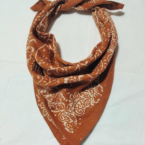 Könnte beinhalten: Ein rostfarbenes Bandana mit weißem Paisley-Muster. Das quadratische Tuch ist gefaltet und geknotet, wodurch das aufwendige Design zur Geltung kommt. Der Stoff ist weich und scheint aus Baumwolle zu sein, geeignet für verschiedene Zwecke.
