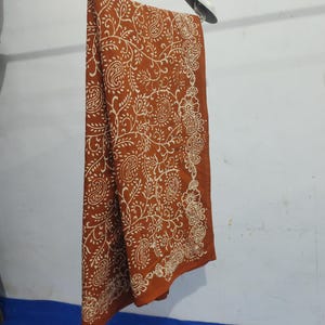 Könnte beinhalten: Ein rostfarbenes Textil mit einem weißen Paisley-Muster wird auf einem Kleiderbügel präsentiert. Der Stoff ist drapiert und zeigt das komplizierte Design. Das Paisley-Muster ist über das gesamte Textil hinweg konsistent, mit einem detaillierten Rand.